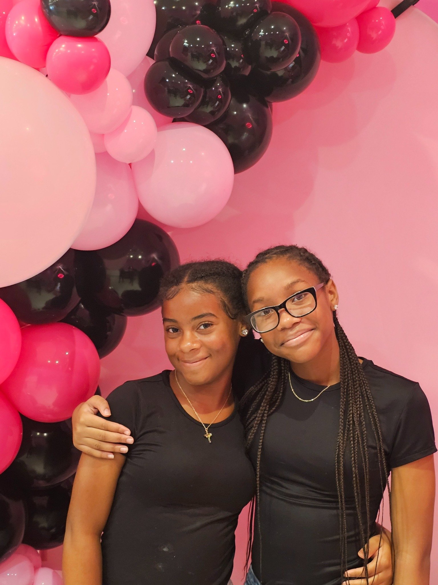 Two young girls standing together in front of a pink backdrop with pink and black balloons