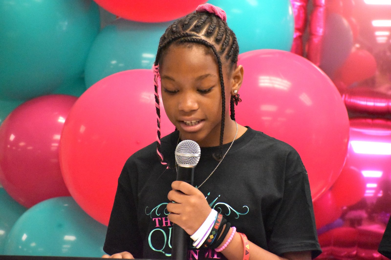 A girl speaking into a microphone at an Enough Period gathering, with pink and teal balloons behind her
